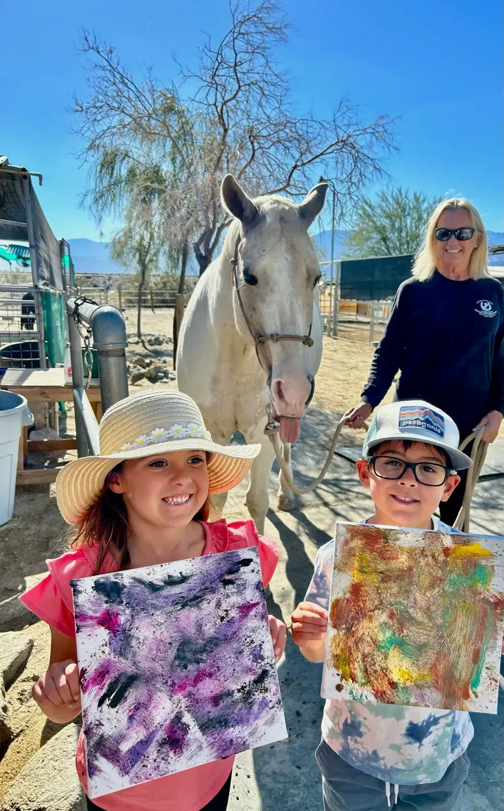 Two children holding paintings they made with a horse, with a volunteer holding the horse nearby